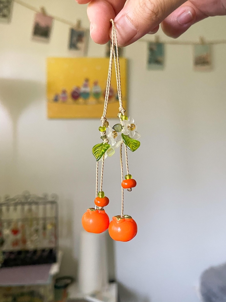 May include: A hanging ornament featuring two bright orange fruit charms suspended from thin beige cords. The design includes small translucent white flower blossoms, green glass leaves, and tiny green and orange accent beads along the strings.