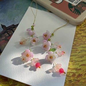 Puede incluir: Primer plano de delicados colgantes florales con pétalos rosas y transparentes, cuentas verdes y una cuerda dorada. Estos accesorios hechos a mano presentan un grupo de flores y una sola cuenta roja.