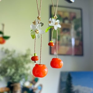 May include: Two hanging ornaments featuring bright orange fruit charms, small green beads, and white flower blossoms with green leaves. The charms are suspended from thin tan cords, creating a delicate, dangling display of botanical-inspired decor.