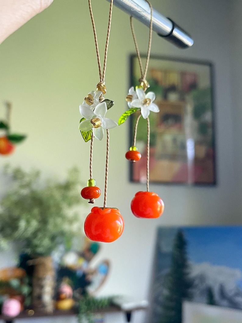 May include: Two hanging ornaments featuring bright orange fruit charms, small green beads, and white flower blossoms with green leaves. The charms are suspended from thin tan cords, creating a delicate, dangling display of botanical-inspired decor.