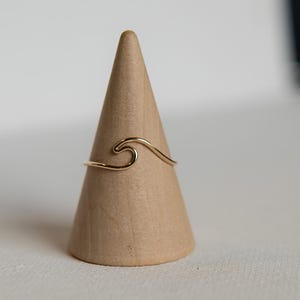 May include: A gold-coloured ring in the shape of a wave. The ring is displayed on a small, light brown, cone-shaped stand. The ring's design is simple and elegant, with a flowing, curved shape.
