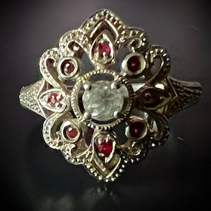 May include: A silver-toned ring with an ornate, floral design. The ring features a large, clear central stone surrounded by smaller red gemstones. The setting has intricate detailing, with a vintage or antique aesthetic.