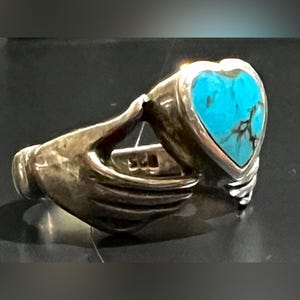 May include: A silver ring featuring a heart-shaped turquoise stone. The ring's design incorporates a hand holding the heart. The ring is marked with "925". The turquoise stone has dark veining.