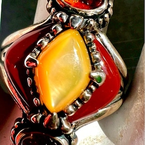 May include: A silver ring with a central yellow gemstone, surrounded by a red, diamond-shaped setting. Two oval, amber-colored stones are set at the top and bottom of the ring. The ring has a detailed, ornate design.