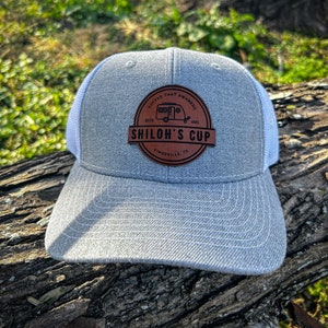 May include: Gray and white trucker hat with a brown leather patch that says "Coffee That Awakens" and "Shiloh's Cup" with a small camper icon and the text "Est. 2023" and "Kingsville, TX".