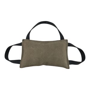 May include: A rectangular, olive-green suede dog toy with black nylon handles. The toy has a stitched border and a loop handle on top, with two additional handles on the sides. The background is plain white.