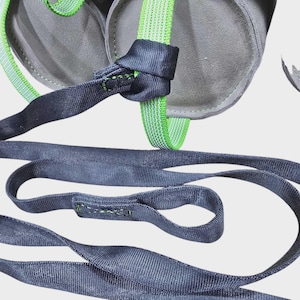 May include: Close-up of a climbing harness with gray padding and black and green straps. The straps are connected with a knot. The harness is designed for safety and support during climbing activities.