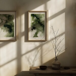 May include: Two framed abstract paintings with green and white watercolor designs hang on a white wall. A vase with a dried branch sits on a wooden table with a small bowl.