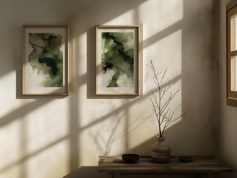 May include: Two framed abstract paintings with green and white watercolor designs hang on a white wall. A vase with a dried branch sits on a wooden table with a small bowl.