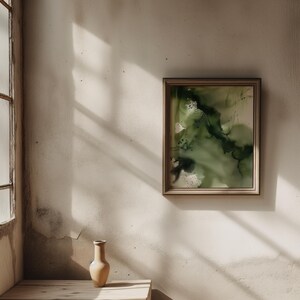 May include: A beige ceramic vase sits on a wooden table in front of a white wall with a framed abstract painting of green and black watercolor.