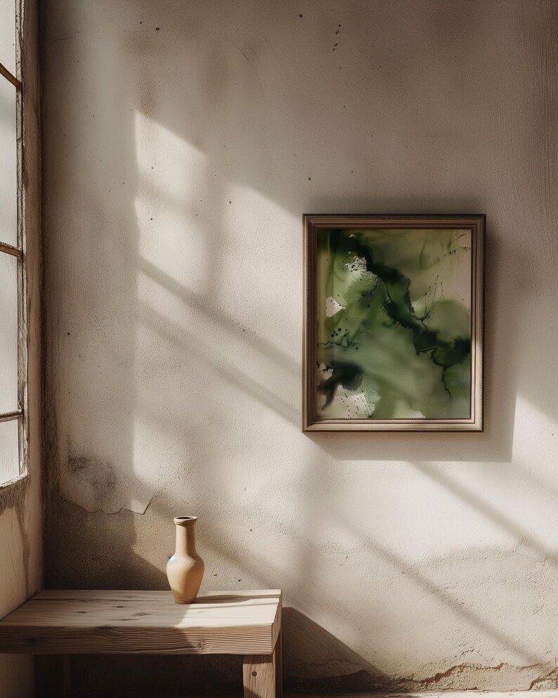 May include: A beige ceramic vase sits on a wooden table in front of a white wall with a framed abstract painting of green and black watercolor.
