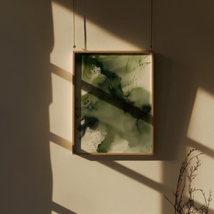 May include: A framed abstract painting with shades of green and white. The painting is hanging on a light beige wall with a shadow from a window.