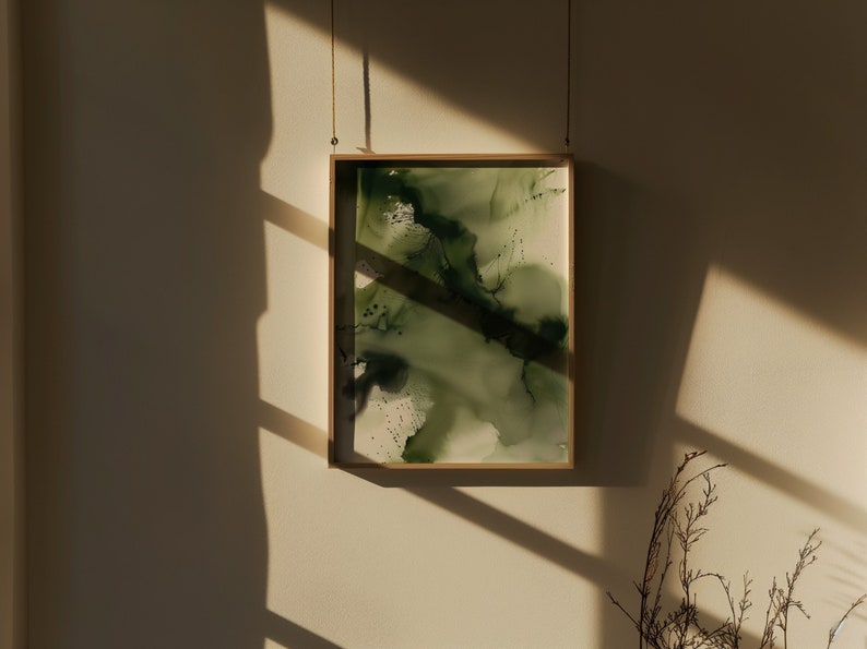 May include: A framed abstract painting with shades of green and white. The painting is hanging on a light beige wall with a shadow from a window.