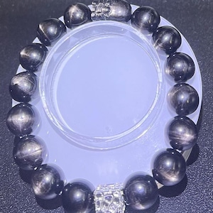 May include: A black gemstone bracelet with silver accents. The bracelet is made of round beads with a shimmering, iridescent finish. The silver accents are in the shape of a square and a small, detailed design.