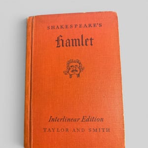 May include: A hardback book with a vibrant orange cloth cover. The front cover features black text reading SHAKESPEARE'S Hamlet, Interlinear Edition, and TAYLOR AND SMITH. A small black graphic of a theatrical mask is centred on the cover.