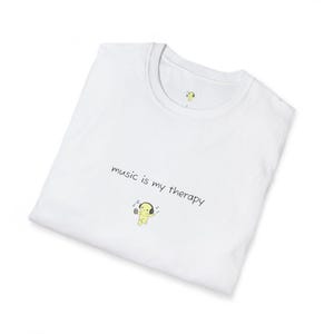 Pode incluir: Camiseta branca com a frase "music is my therapy" em escrita preta. Uma pequena figura de desenho animado amarela usando fones de ouvido é retratada abaixo do texto, com notas musicais ao redor. A camisa tem gola redonda.