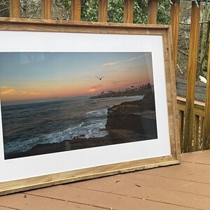 May include: A framed photograph of a sunset over the ocean with a bird flying in the sky. The frame is made of wood and has a white mat.