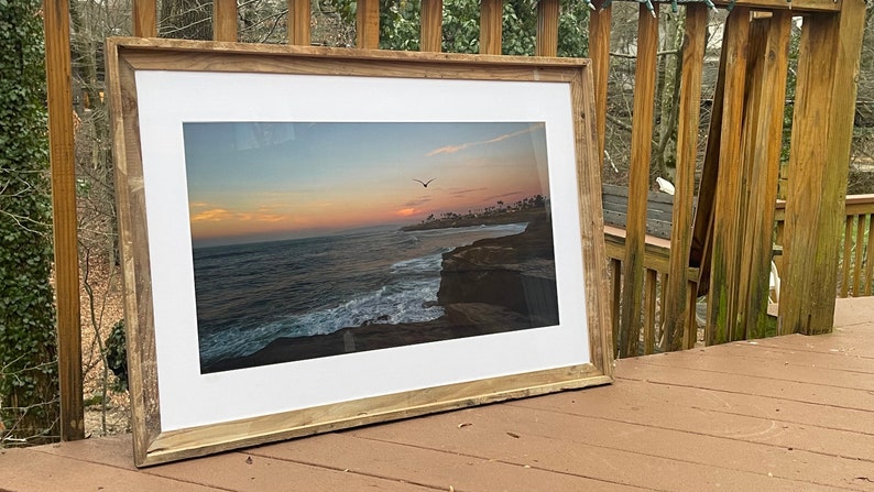 May include: A framed photograph of a sunset over the ocean with a bird flying in the sky. The frame is made of wood and has a white mat.