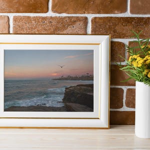 May include: A white picture frame with a gold trim, displaying a photograph of a sunset over the ocean with a bird flying in the sky. The frame is sitting on a wooden surface in front of a brick wall.