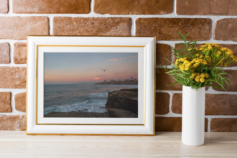 May include: A white picture frame with a gold trim, displaying a photograph of a sunset over the ocean with a bird flying in the sky. The frame is sitting on a wooden surface in front of a brick wall.