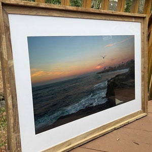 May include: A framed photograph of a sunset over the ocean with a bird flying in the sky. The frame is made of wood and has a white mat.
