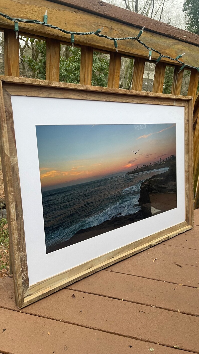 May include: A framed photograph of a sunset over the ocean with a bird flying in the sky. The frame is made of wood and has a white mat.