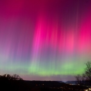 May include: A vibrant display of the aurora borealis, also known as the Northern Lights, with shades of pink and green illuminating the night sky. The lights are visible above a dark silhouette of a forest and a distant town.