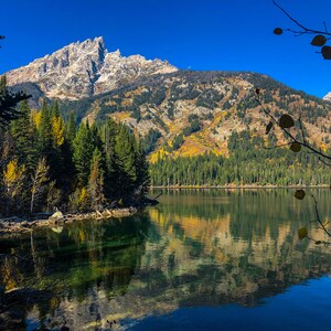 May include: A scenic view of a mountain range reflected in a still lake. The mountains are covered in snow and trees, and the lake is surrounded by a forest. The sky is blue and clear.