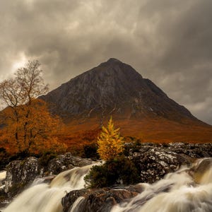 May include: A cascading waterfall flows over moss-covered rocks in front of a mountain with a cloudy sky. The water is white and the rocks are gray. A small yellow tree grows on the right side of the image.