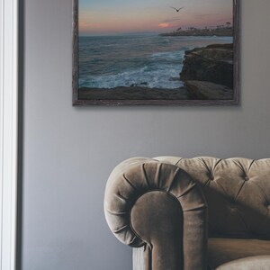 May include: A framed photograph of a seascape with a bird flying over the ocean. The sky is a soft pink and blue, and the waves are crashing against the rocky shore.