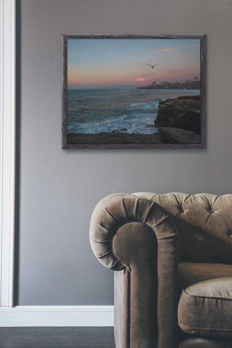 May include: A framed photograph of a seascape with a bird flying over the ocean. The sky is a soft pink and blue, and the waves are crashing against the rocky shore.