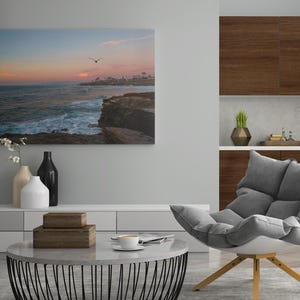 May include: A living room interior design with a gray armchair, a coffee table, and a framed photograph of a sunset over the ocean. The photograph is hanging on a light gray wall. The armchair is positioned in front of the coffee table, which is made of white marble with a black metal base. The coffee table is decorated with two wooden boxes and a cup of coffee. The armchair is upholstered in gray fabric and has a white base. The room is decorated with a vase of white flowers and a black vase.