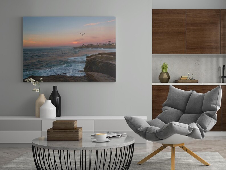 May include: A living room interior design with a gray armchair, a coffee table, and a framed photograph of a sunset over the ocean. The photograph is hanging on a light gray wall. The armchair is positioned in front of the coffee table, which is made of white marble with a black metal base. The coffee table is decorated with two wooden boxes and a cup of coffee. The armchair is upholstered in gray fabric and has a white base. The room is decorated with a vase of white flowers and a black vase.