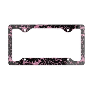 May include: Black license plate frame with a pink lace pattern. The frame has four mounting holes and a rectangular shape. The lace design is intricate and covers the entire frame.