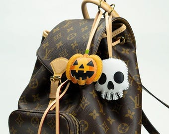 May include: A brown monogram print leather backpack featuring two hanging charms: an orange and black leather pumpkin jack-o'-lantern and a white and black leather skull, both attached to the top handle with light tan leather straps.