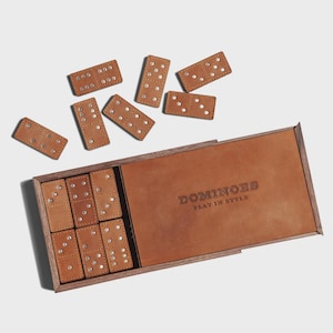 Può includere: Un set di domino in pelle marrone con segni a punti argentati. Il set include una scatola di legno con un coperchio in pelle e il testo "DOMINOES PLAY IN STYLE" in rilievo sul coperchio. Diversi domino sono sparsi intorno alla scatola.