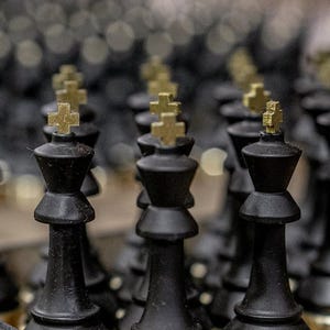 May include: Close-up of black chess pieces with gold-colored crosses on top. The pieces are arranged in rows, with the focus on the kings. The background is blurred, creating a shallow depth of field. The chess pieces are made of a dark material.