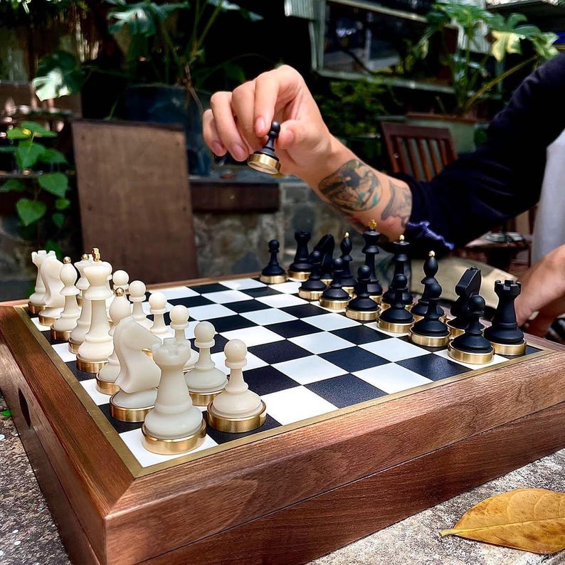 May include: A wooden chess set with black and white checkered board. The chess pieces are black and off-white with gold-colored bases. A hand is shown moving a black pawn. The set is on a wooden surface.