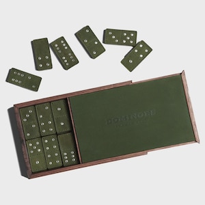 May include: A set of olive green dominoes with silver dot markings, displayed with a wooden box. The box has a dark green interior and the word "DOMINOES" embossed on the lid. The dominoes are scattered around the open box.