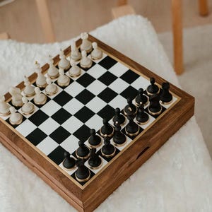 May include: A wooden chess set with black and white pieces arranged on a checkered board. The chess pieces are black and white with gold accents. The board is set inside a wooden box with a gold border.