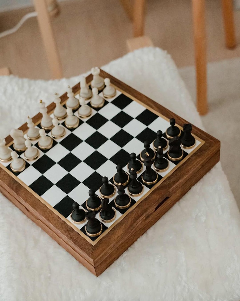 May include: A wooden chess set with black and white pieces arranged on a checkered board. The chess pieces are black and white with gold accents. The board is set inside a wooden box with a gold border.