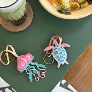 May include: Two leather keychains featuring a pink and blue jellyfish and a blue and pink turtle. The keychains are on a green surface.