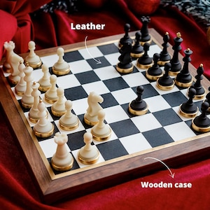 May include: A chess set with a wooden case and a black and white checkered board. The chess pieces are ivory and black with gold-colored bases. The image shows the pieces arranged on the board, ready for a game.
