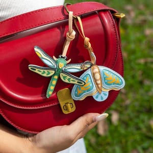 May include: A red leather handbag with a gold clasp features two leather charms: a green and yellow dragonfly and a blue and yellow moth. The charms are attached with tan leather straps.