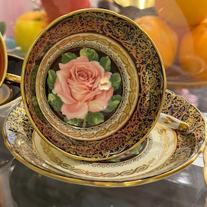 Può includere: Un set vintage di tazza da tè e piattino con un design floreale dorato e blu. La tazza presenta una rosa rosa con foglie verdi e un bordo dorato. Il piattino ha un design abbinato.