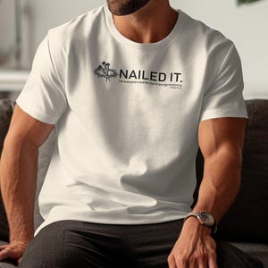 Puede incluir: Camiseta blanca de cuello redondo con el texto negro "NAILED IT.", un pequeño gráfico y texto adicional. La camiseta está hecha de un material suave.