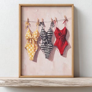 Op de afbeelding: Een ingelijste print met drie badpakken die aan een waslijn hangen. De badpakken zijn geel met witte stippen, zwart-wit gestreept en effen rood. De afbeelding heeft een lichtroze achtergrond.