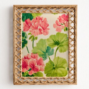 May include: A framed floral print featuring pink geraniums and large green leaves. The artwork is set within a rectangular, woven, light brown frame. The background is a cream color, and the overall style is reminiscent of a watercolor painting.