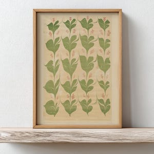 May include: A framed print featuring a repeating pattern of green leaves and small pink flowers on a beige background. The artwork is displayed in a light brown wooden frame and rests on a wooden shelf.