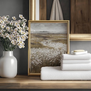 May include: A still-life arrangement featuring a white vase with white flowers, a framed landscape painting, and a stack of white towels. The painting has a gold frame and depicts a field of white flowers. A small white soap dish with a bar of soap is on top of the towels.